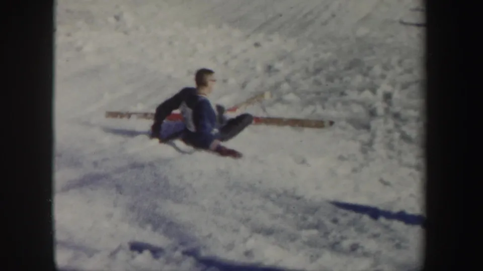 The skier sits on the snow with skis spread behind him after the slip, framed from a spectator viewpoint on the New York slope.