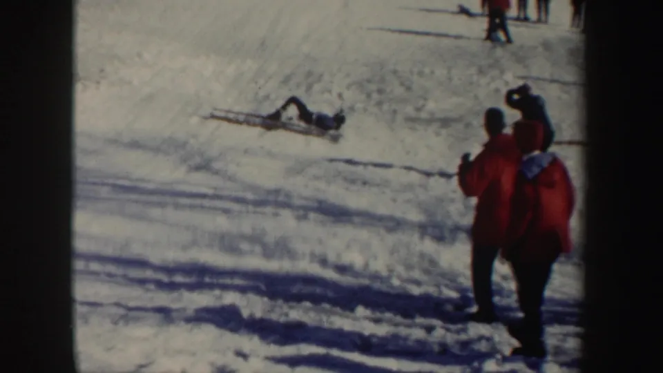 A skier lies on the snowy landing area as spectators in red winter coats watch from the side of the 1962 New York ski slope.