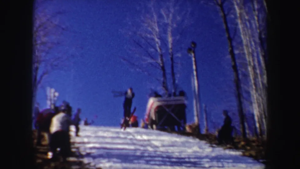 A skier in a white sweater is seen on the 1962 New York ski slope near a jump or landing area, with spectators and bare trees nearby.
