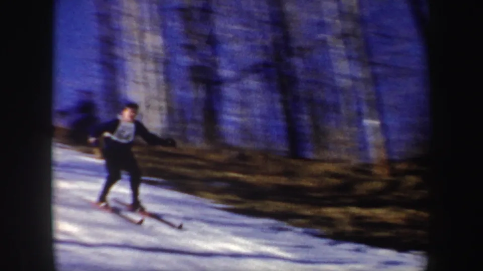 1962 New York 8mm frame of a skier in dark clothing descending a snowy slope near bare trees.
