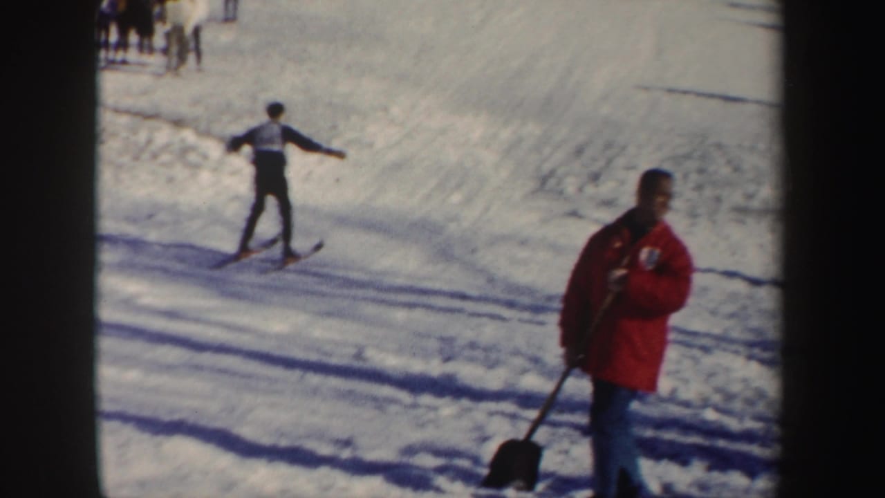 1962 New York ski-slope frame with a skier on the course and a red-jacketed person standing with a shovel.