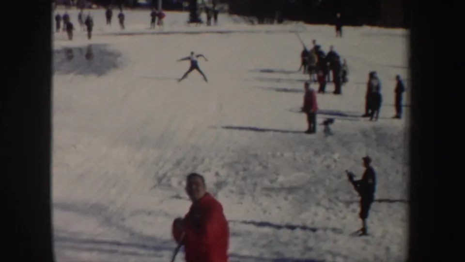 Wide 1962 New York 8mm frame showing a skier on the slope, clustered spectators, and a red-jacketed figure in front.