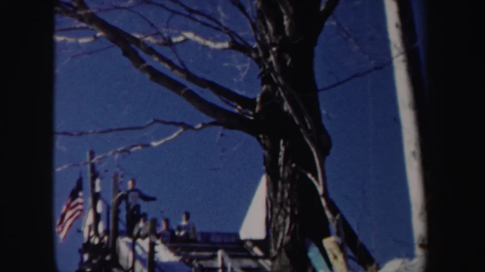 1962 New York ski-area scene with people near a snowy rise, an American flag, bare branches, and a deep blue sky.