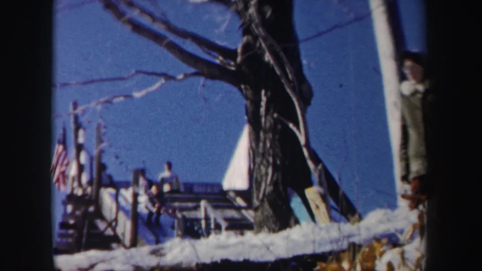 A 1962 New York home-movie frame shows people near a snowy ski slope, with a flag, bare trees, and a figure partly cut off at the edge.