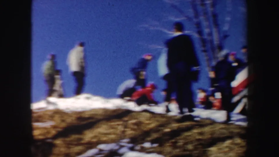 1962 New York winter recreation frame with people on a snowy rise, including a bent or fallen figure, under clear blue sky.