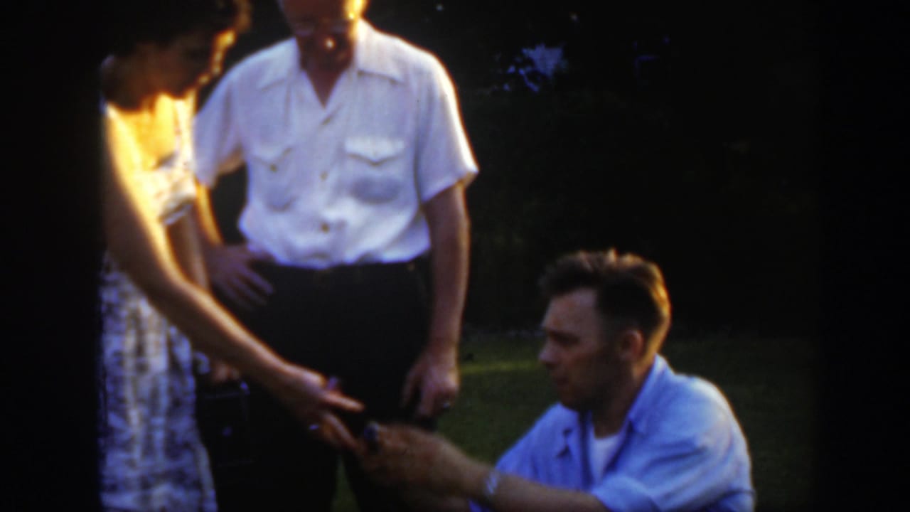 In supported 1949 Massachusetts home-movie footage, a man kneels before a woman on a lawn as another man stands close by.