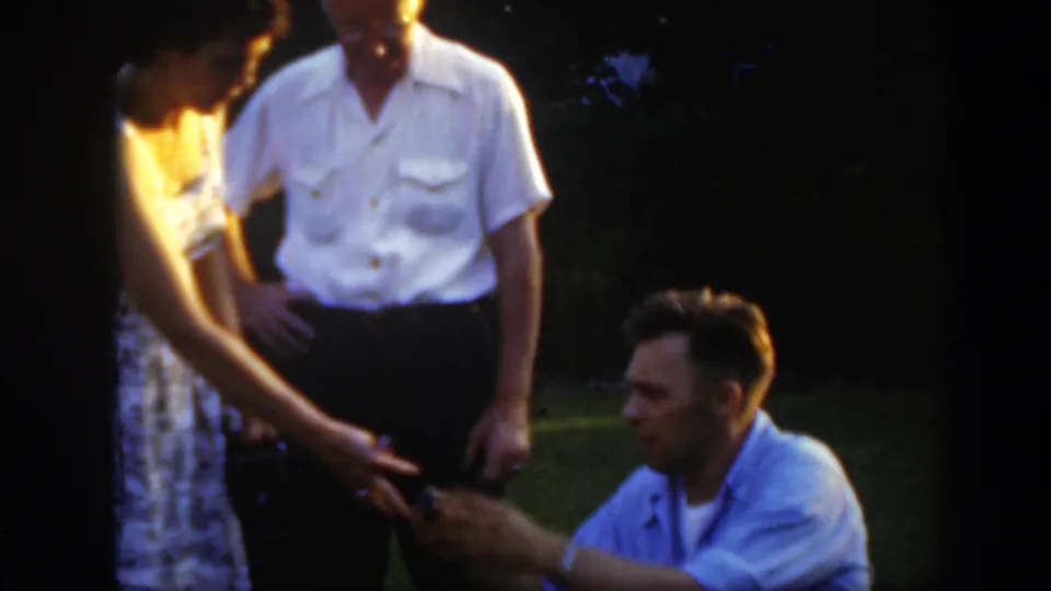 In supported 1949 Massachusetts home-movie footage, a man kneels before a woman on a lawn as another man stands close by.