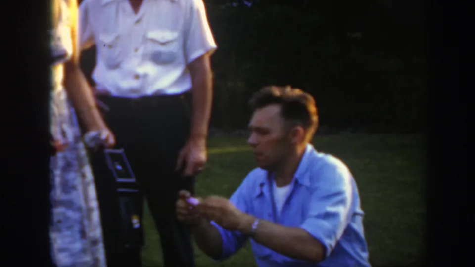 A kneeling man in a light blue shirt handles a small object as adults stand nearby in a Massachusetts lawn setting.