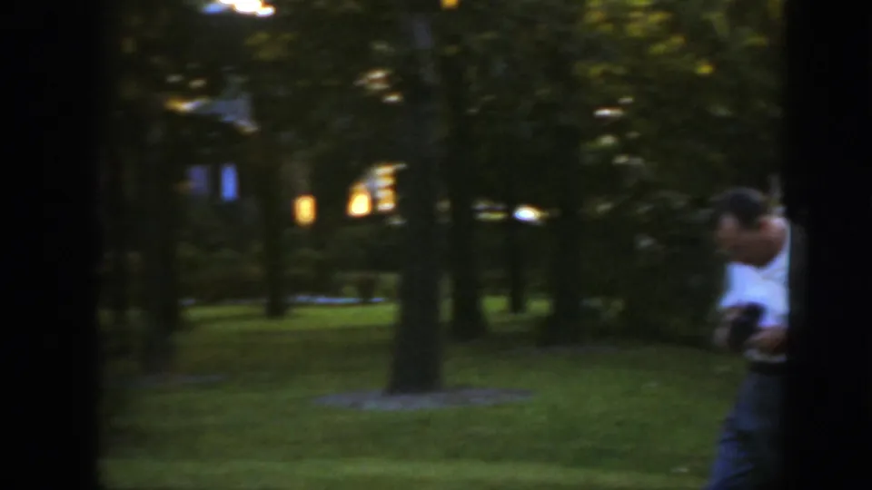 A man moves across a shaded Massachusetts lawn, looking down at a small item or camera in his hands.
