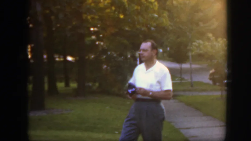 An adult man stands in warm evening light on a Massachusetts lawn near a sidewalk, holding a small dark object.