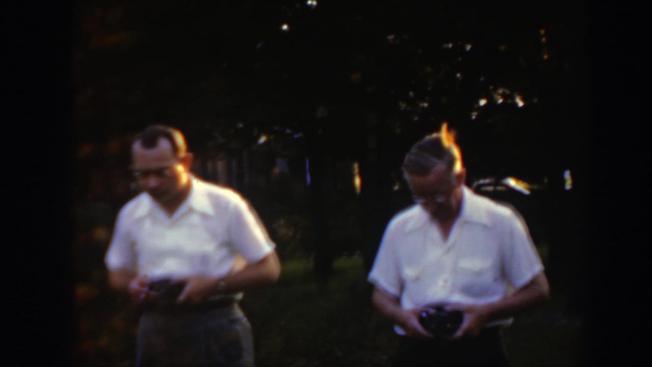 Two men in white shirts stand in a dim yard holding small dark devices.