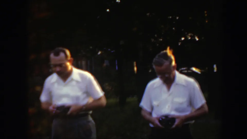 In 1949 Massachusetts home-movie footage, two men stand in a grassy yard at dusk while examining small dark handheld devices.