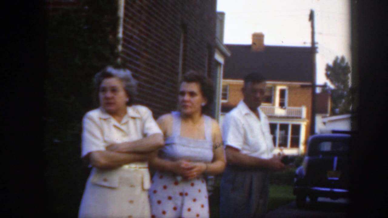 Adults stand near a brick house in Massachusetts, with two women in dresses and a man in a white shirt beside a vintage parked car.