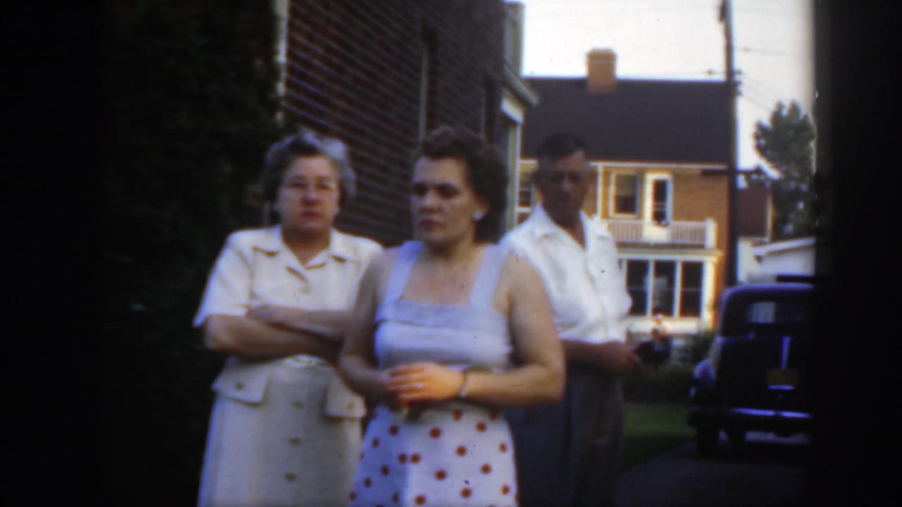 Three adults in late-1940s clothing stand beside a brick residential building, with a dark vintage car and another house in the background.