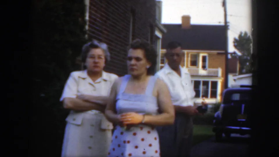 Three adults in late-1940s clothing stand beside a brick residential building, with a dark vintage car and another house in the background.