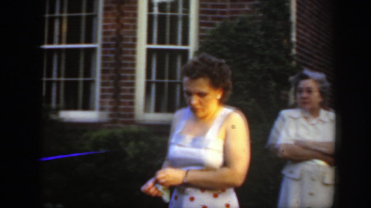 A woman in a light top and polka-dot skirt looks down near a brick house, while another woman stands farther back by the wall.