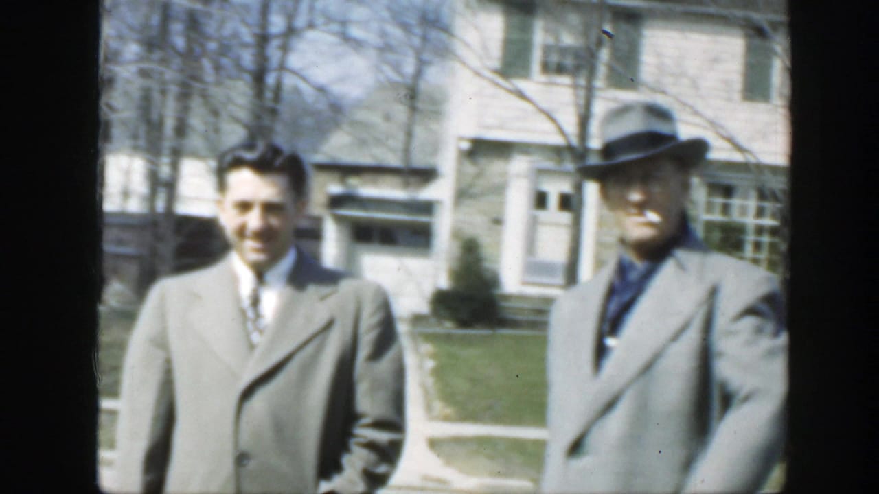 Two men in gray coats stand outside a pale house on a sunny day.