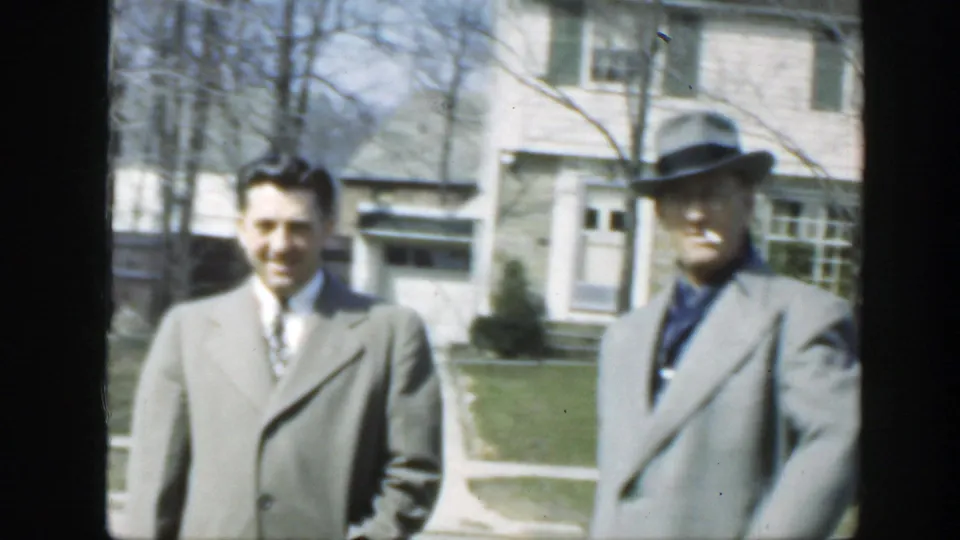 Two adult men stand near a pale house in Massachusetts home-movie footage from 1949; one wears a tie and the other wears a fedora.