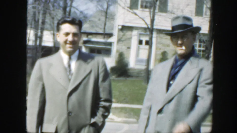 Two adult men remain facing the camera outside a Massachusetts house in 1949, with the older man in a fedora and the younger man in a tie.
