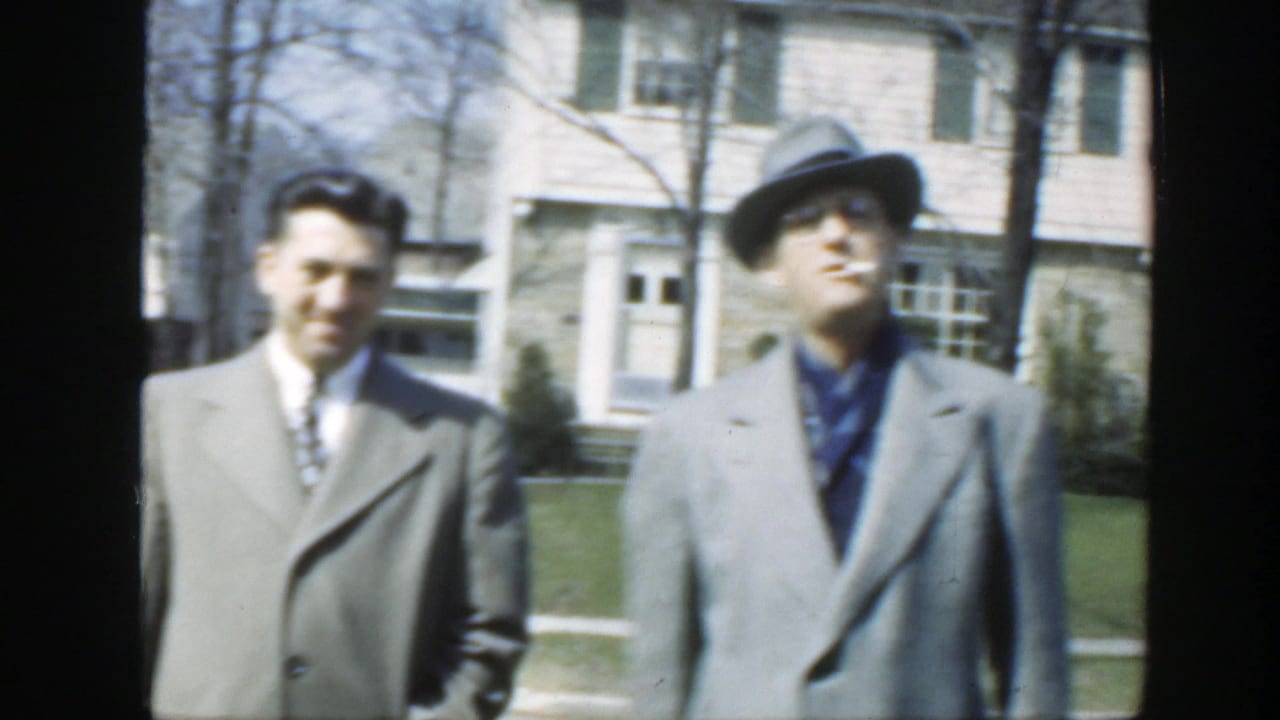 Two adult men stand in bright daylight outside a Massachusetts house in 1949, with the fedora-wearing man slightly turned toward the camera.