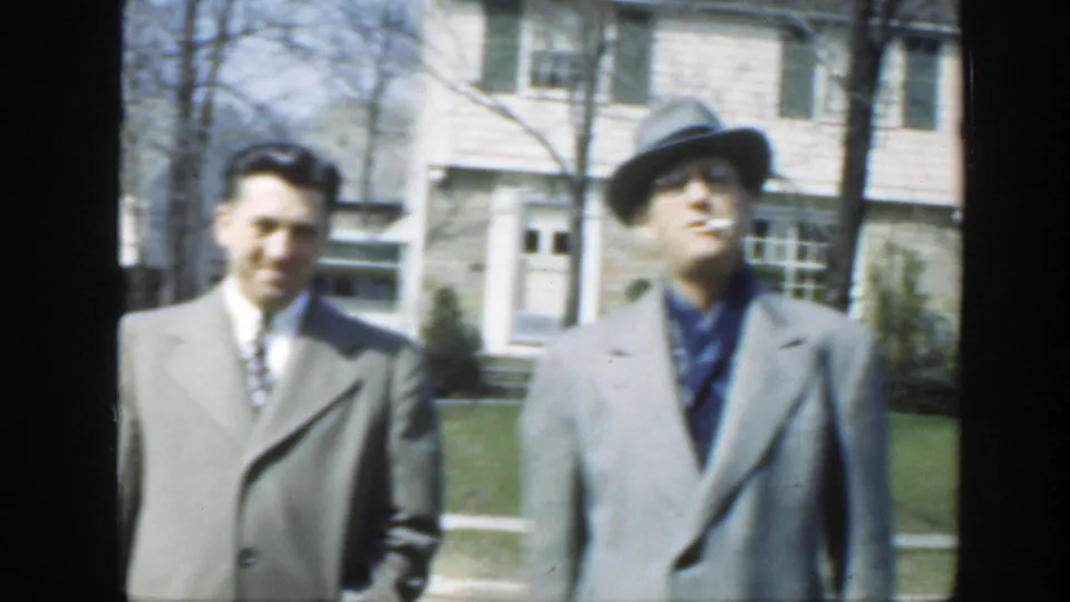 Two adult men stand in bright daylight outside a Massachusetts house in 1949, with the fedora-wearing man slightly turned toward the camera.