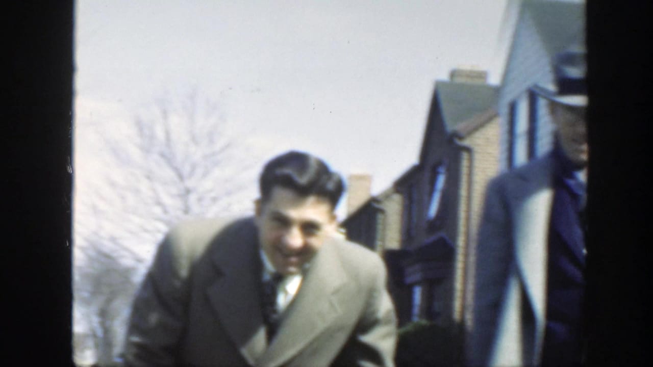 A suited man smiles and leans toward the camera while another suited man in a fedora stands partly at the edge of the frame.