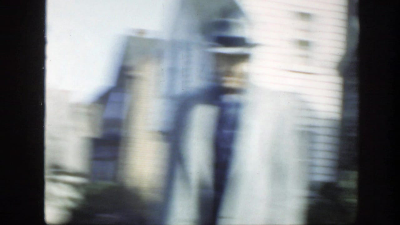 The camera swings past suited figures and a suburban house, producing strong motion blur in the 8mm frame.