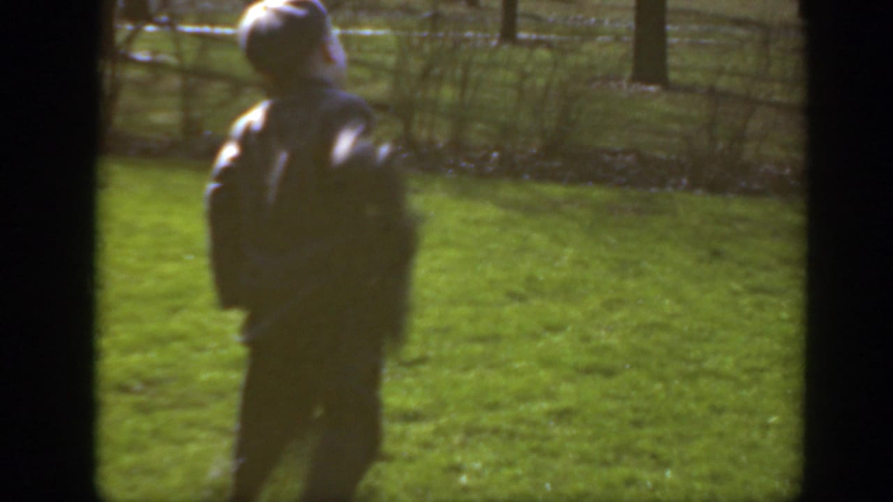 A figure walks across a green suburban yard in Massachusetts home-movie footage, with trees and a fence in the background.
