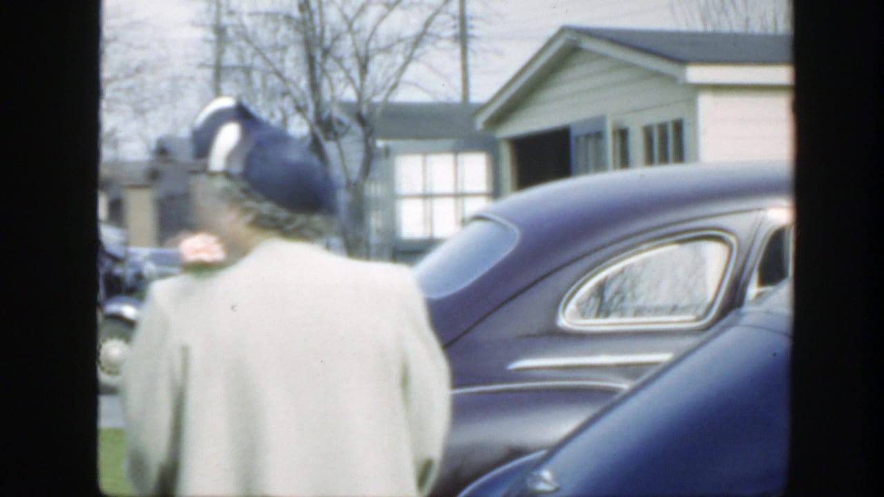 Older woman in a light coat and blue hat walks beside a dark vintage car on a suburban street.