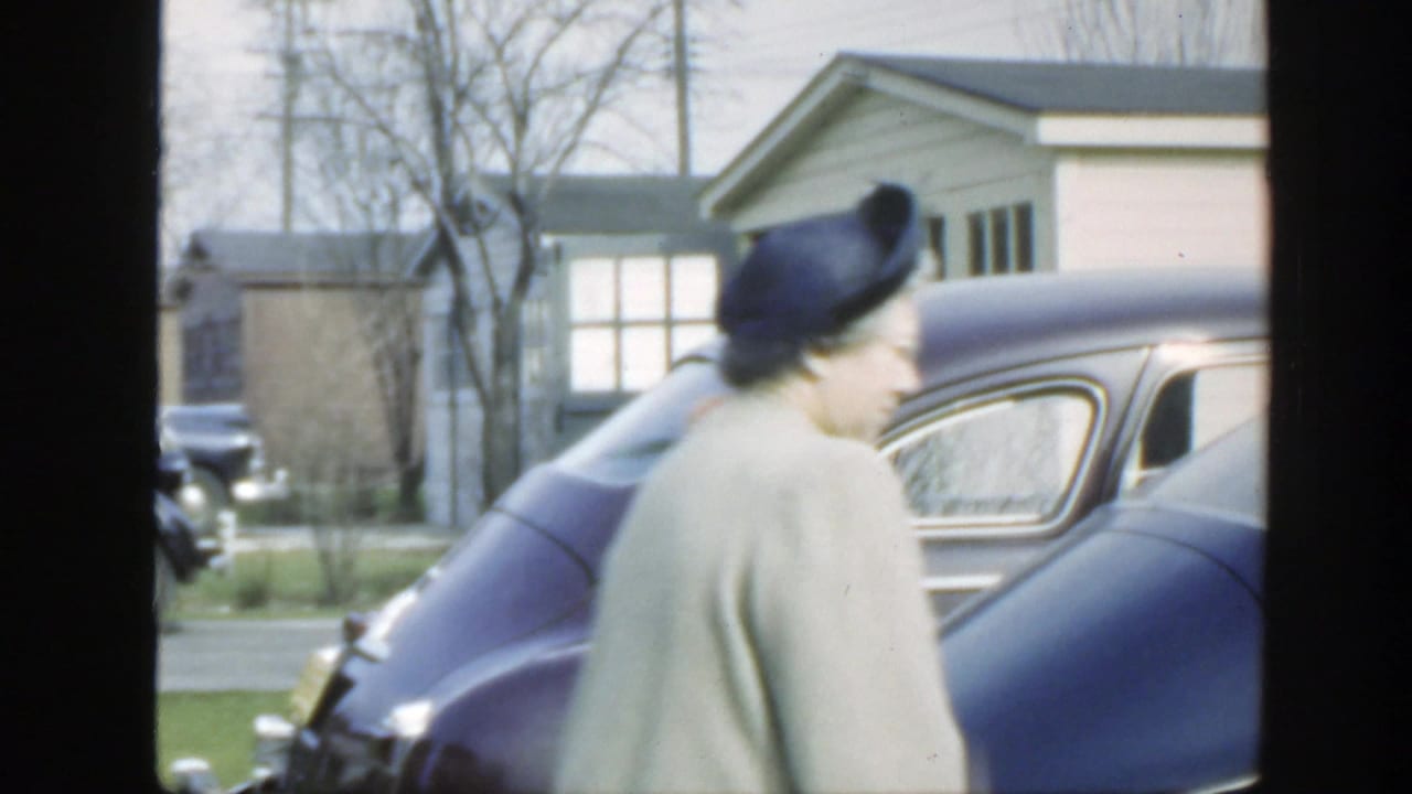 The woman moves past the rear side of a dark purple car in a Massachusetts suburban setting, with bare trees and a small house visible.