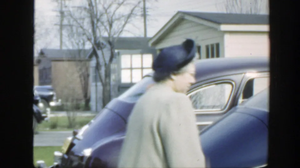 The woman moves past the rear side of a dark purple car in a Massachusetts suburban setting, with bare trees and a small house visible.