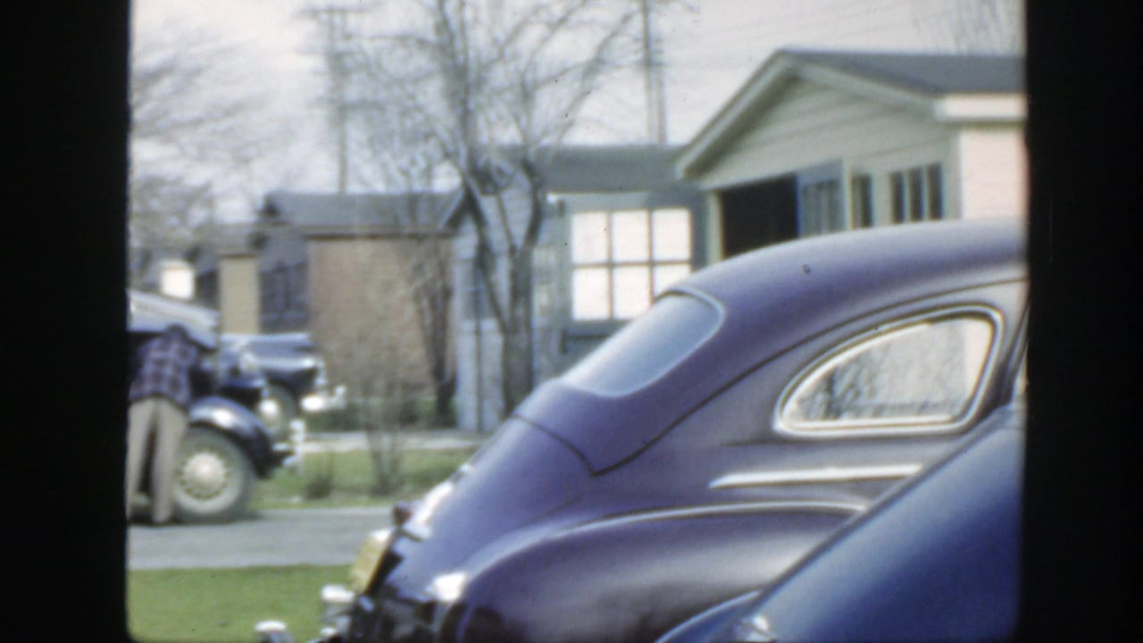 A dark purple car fills the foreground while a man in a plaid shirt stands near another classic car on the residential street.