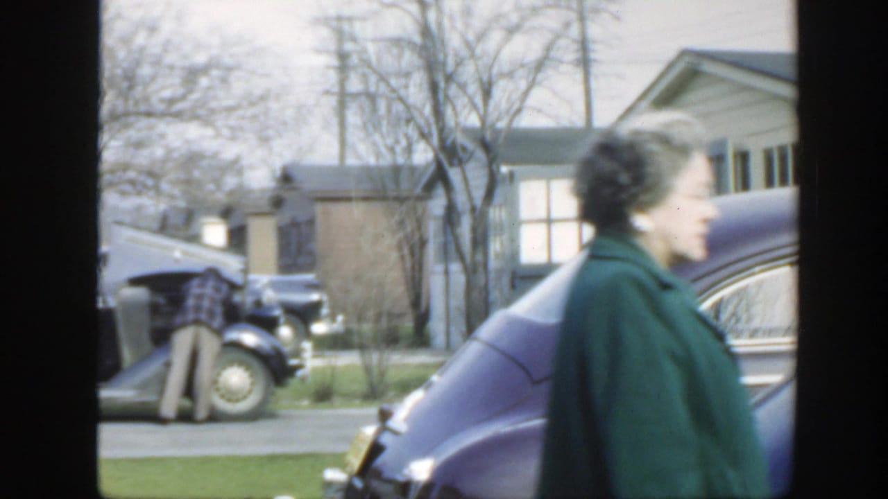 Woman walking past parked cars while a man works under an open car hood.