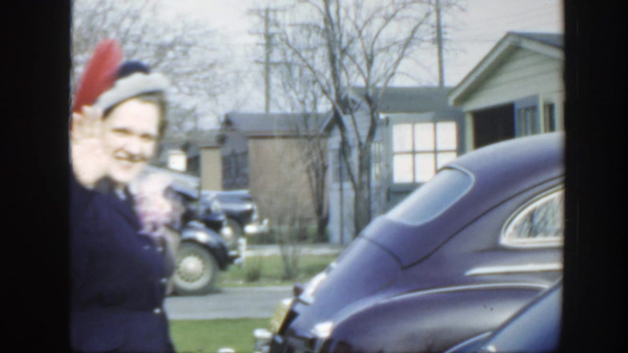1949 Massachusetts frame with a smiling woman in a dark coat and feathered hat walking past the camera beside rounded parked cars.