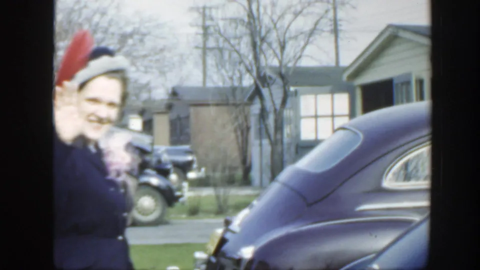 1949 Massachusetts frame with a smiling woman in a dark coat and feathered hat walking past the camera beside rounded parked cars.