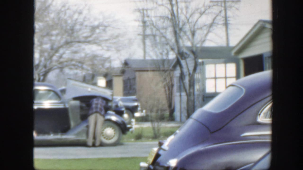 1949 Massachusetts home-movie frame focused on an open car hood, parked cars, houses, lawns, and bare trees on a quiet street.