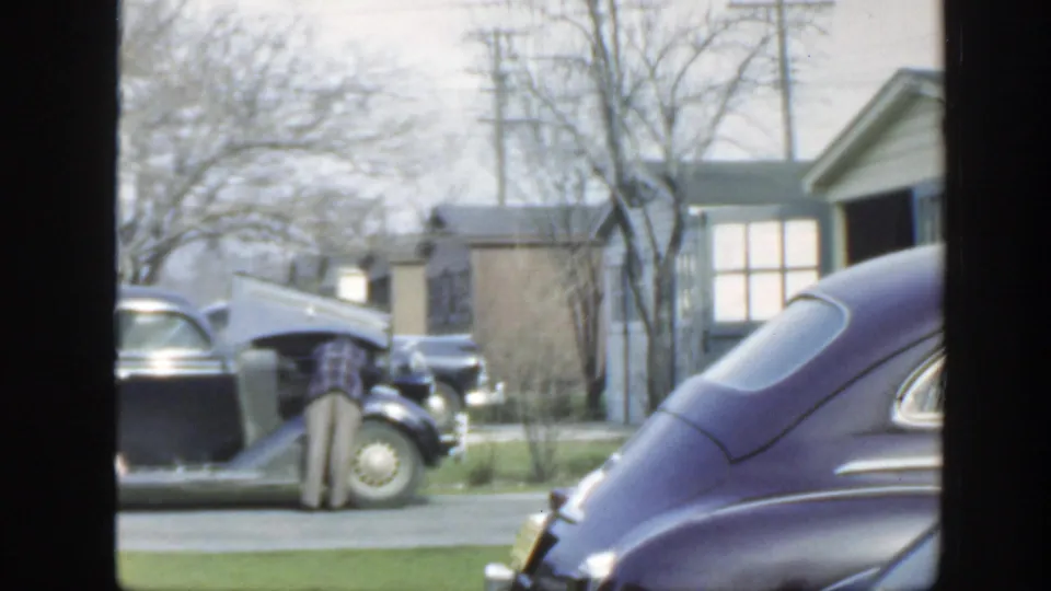 1949 Massachusetts home-movie frame focused on an open car hood, parked cars, houses, lawns, and bare trees on a quiet street.