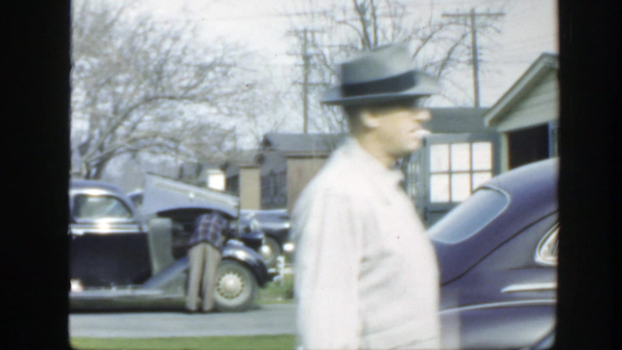 1949 Massachusetts frame showing a man in a light shirt and fedora walking past a dark parked car and open hood on a suburban street.