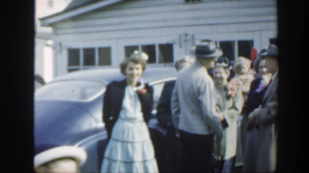 Wedding guests stand outside beside a rounded blue sedan and residential garage.
