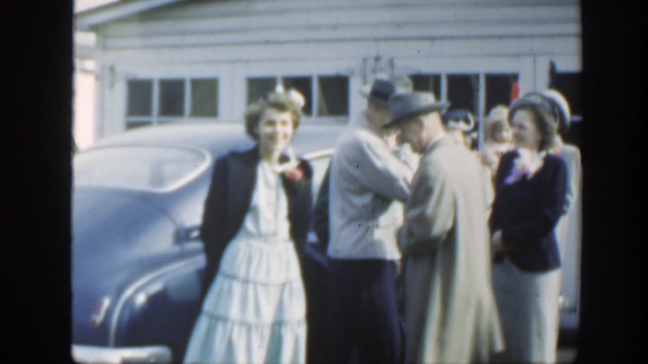 A woman in a white dress and dark jacket stands near a blue sedan as formally dressed guests converse in Massachusetts, 1949.