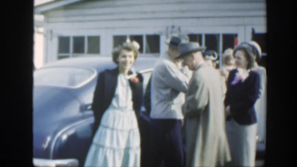 A woman in a white dress and dark jacket stands near a blue sedan as formally dressed guests converse in Massachusetts, 1949.