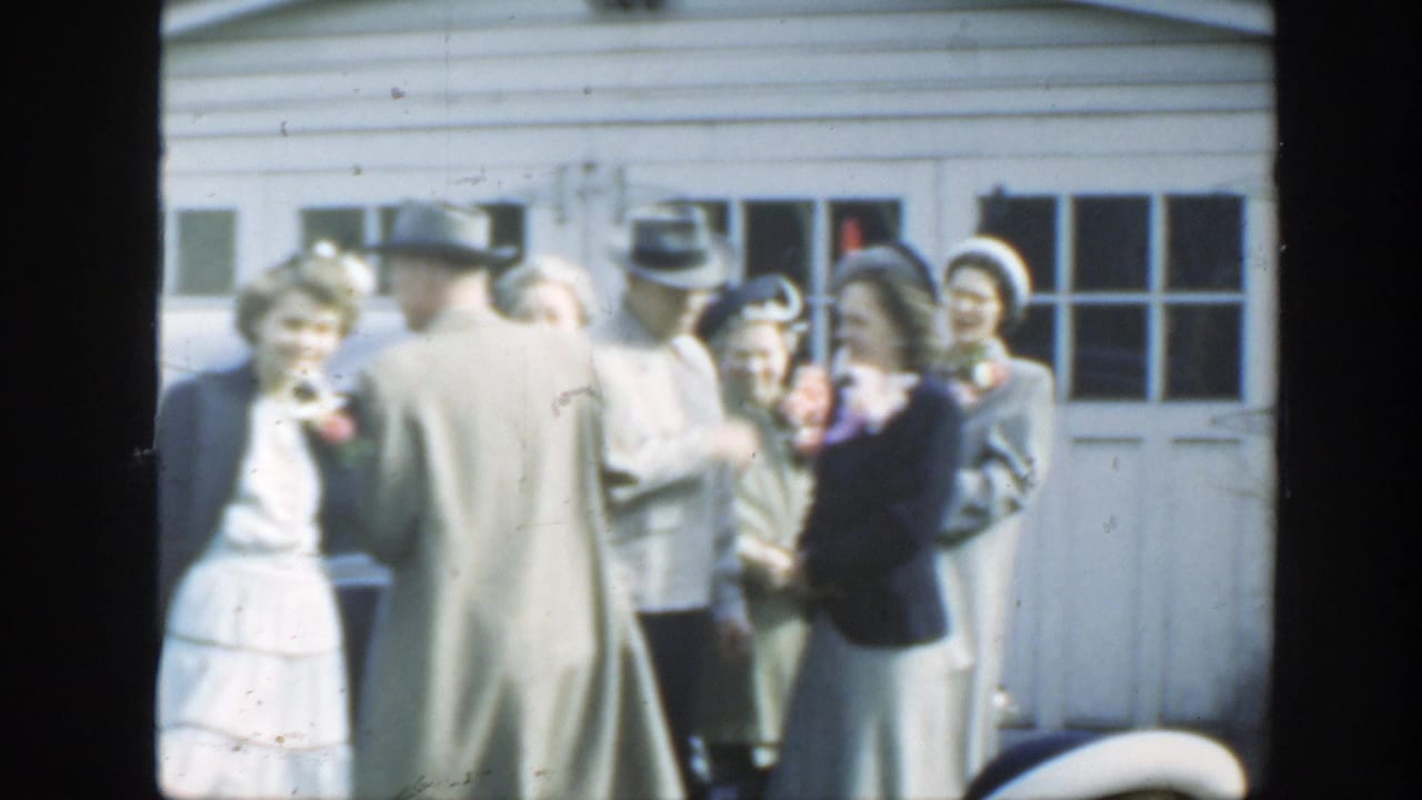 Home-movie frame of 1949 Massachusetts wedding guests in hats and coats gathered outside a residential garage near a parked car.