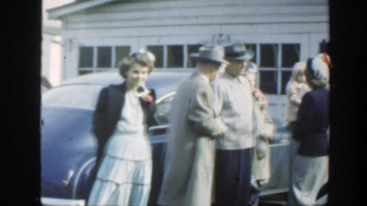 1949 Massachusetts wedding gathering with a woman in white, men in hats, and guests beside a blue sedan outside a garage.