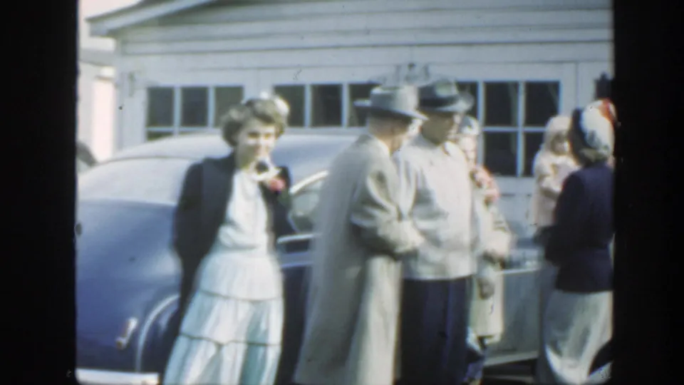 1949 Massachusetts wedding gathering with a woman in white, men in hats, and guests beside a blue sedan outside a garage.