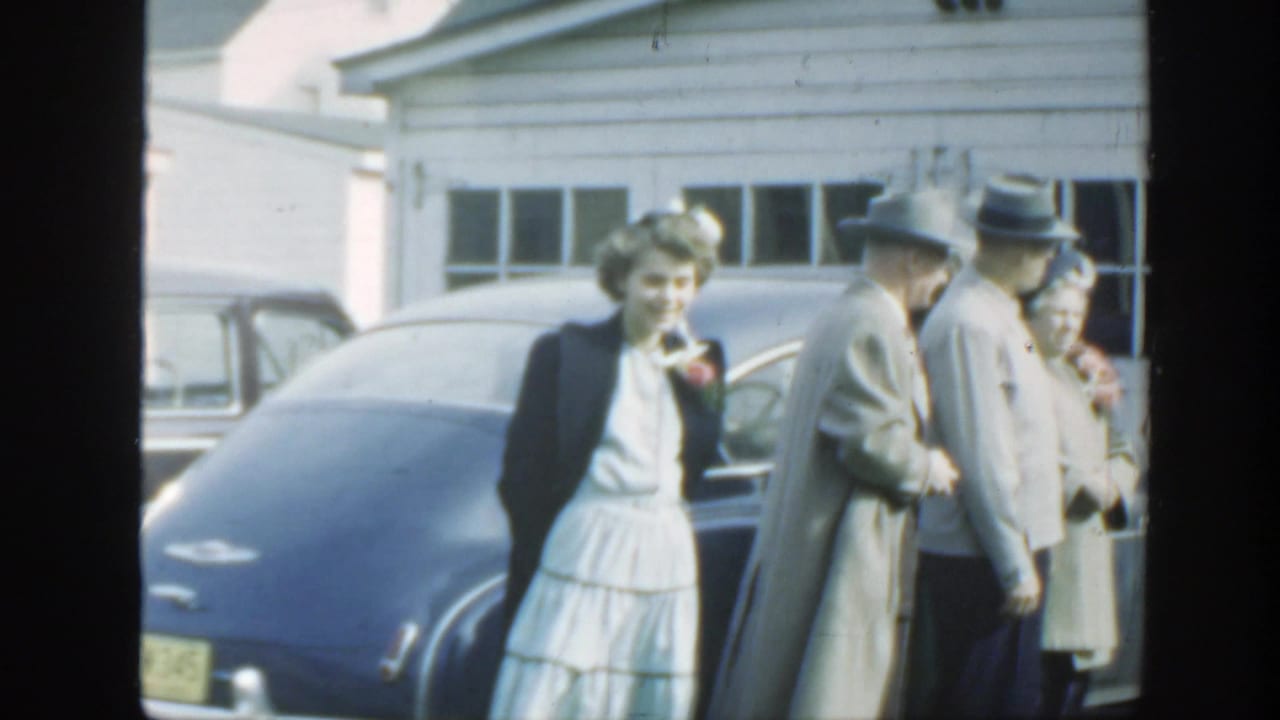 Adults in dress clothes stand beside a rounded blue car near a light-colored house.