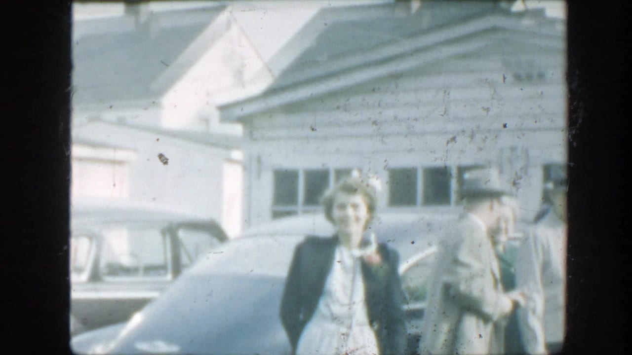 1949 Massachusetts frame with a woman facing the camera and suited adults near parked cars outside a light clapboard house.