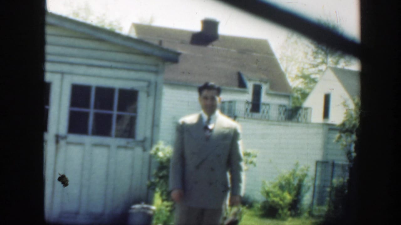 1949 Massachusetts frame of a man in a light double-breasted suit standing in a grassy backyard beside a white shed and houses.