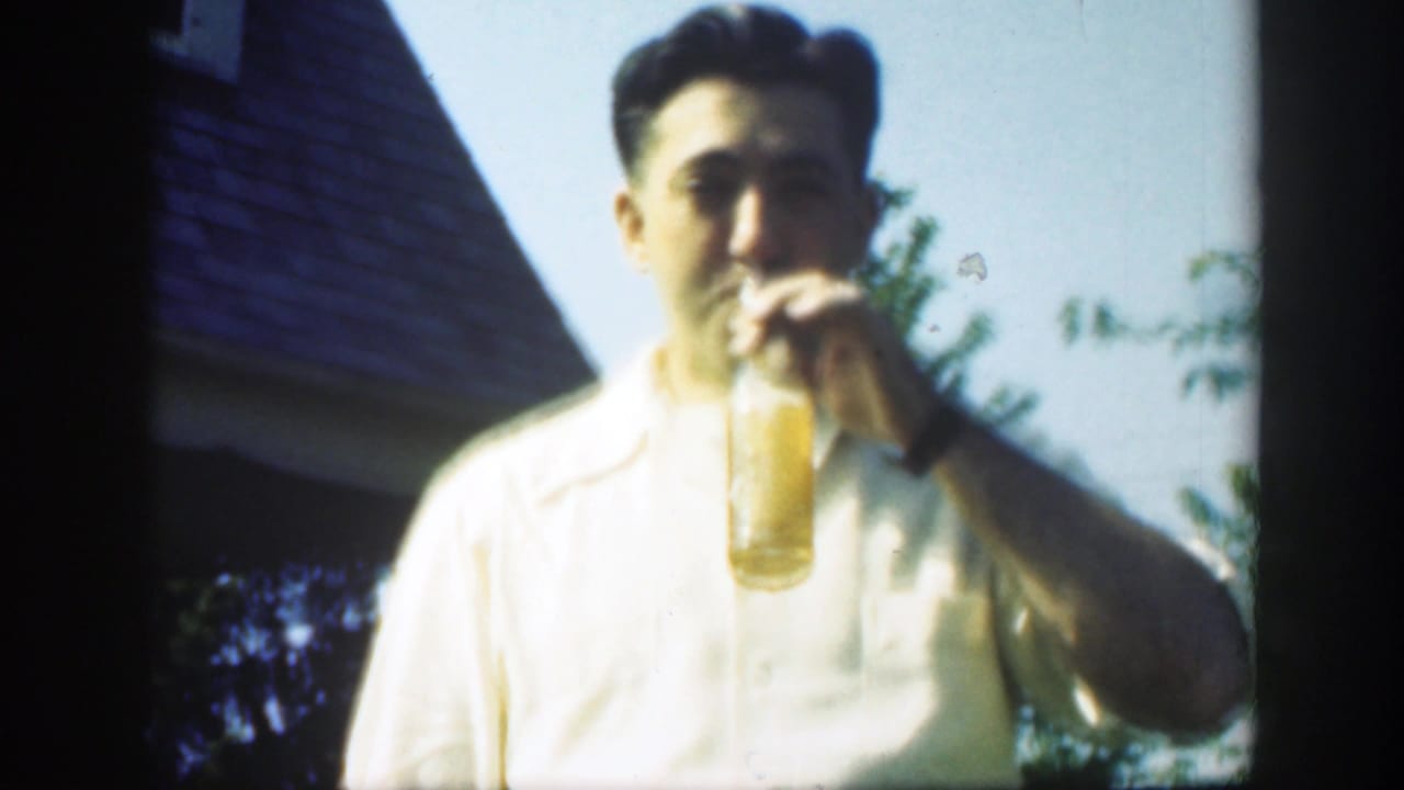 Man in a white short-sleeved shirt drinks from a glass bottle outdoors.