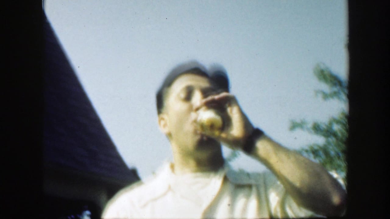 The man raises the bottle higher while drinking, framed against sky, roofline, and yard greenery in the home-movie footage.