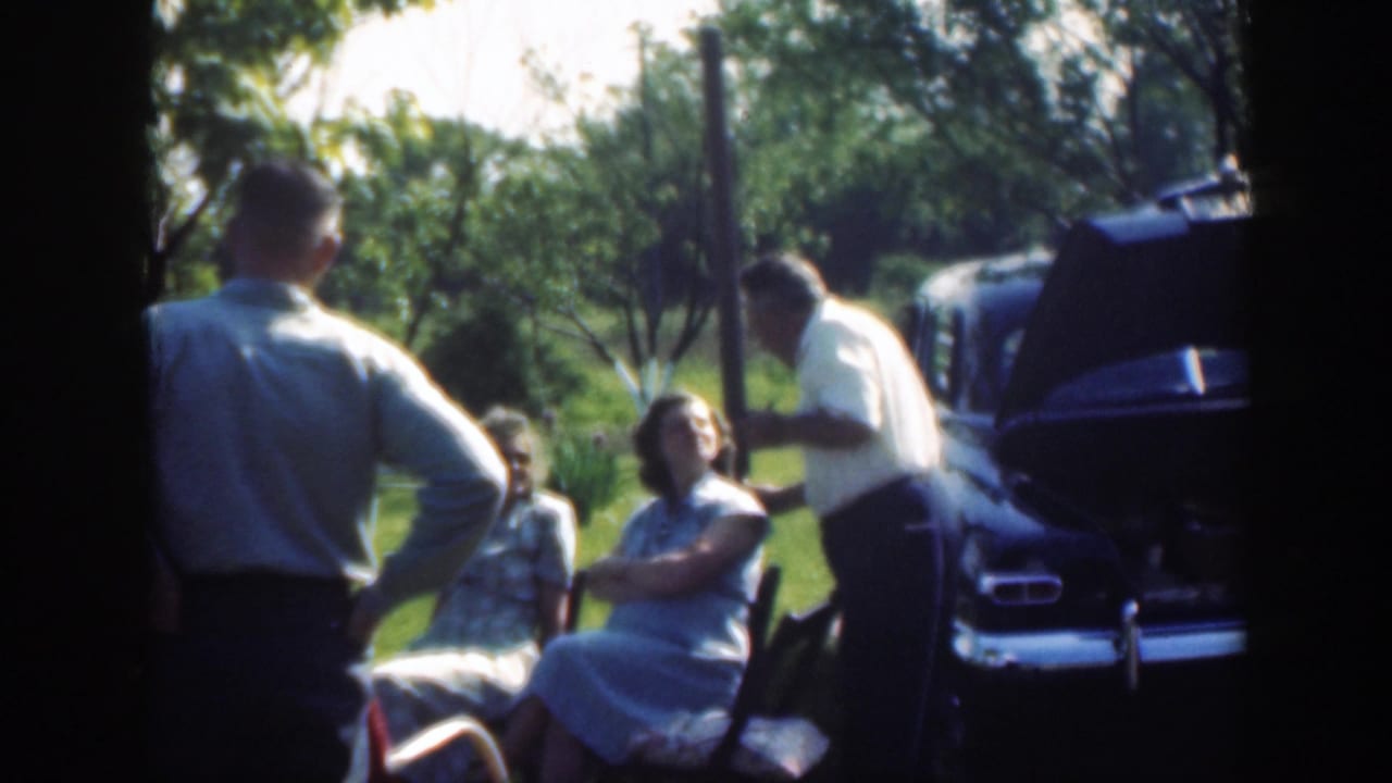 Adults stand and sit near a dark vintage car with its hood open during the 1949 Massachusetts outdoor gathering.